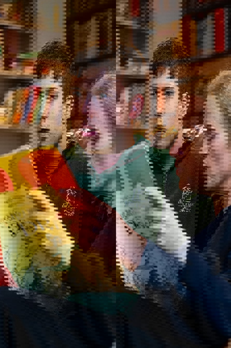 Young person and adult carer sitting together on a sofa, reading a large illustrated book in a cosy room with bookshelves in the background.