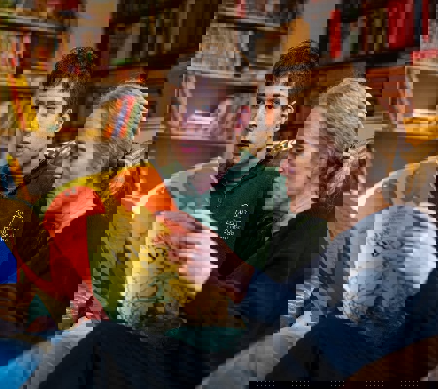 Young person and adult carer sitting together on a sofa, reading a large illustrated book in a cosy room with bookshelves in the background.