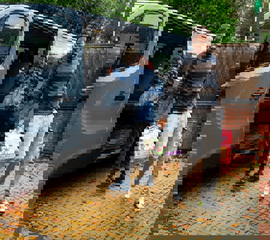 Carer closing the door of a van while a young man carries shopping bags across a driveway.