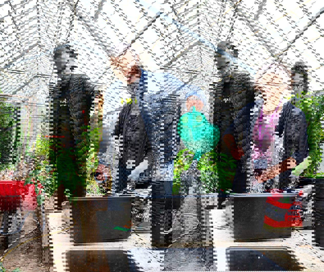 Young man watering plants in a greenhouse while a staff member supports him beside a potting table.