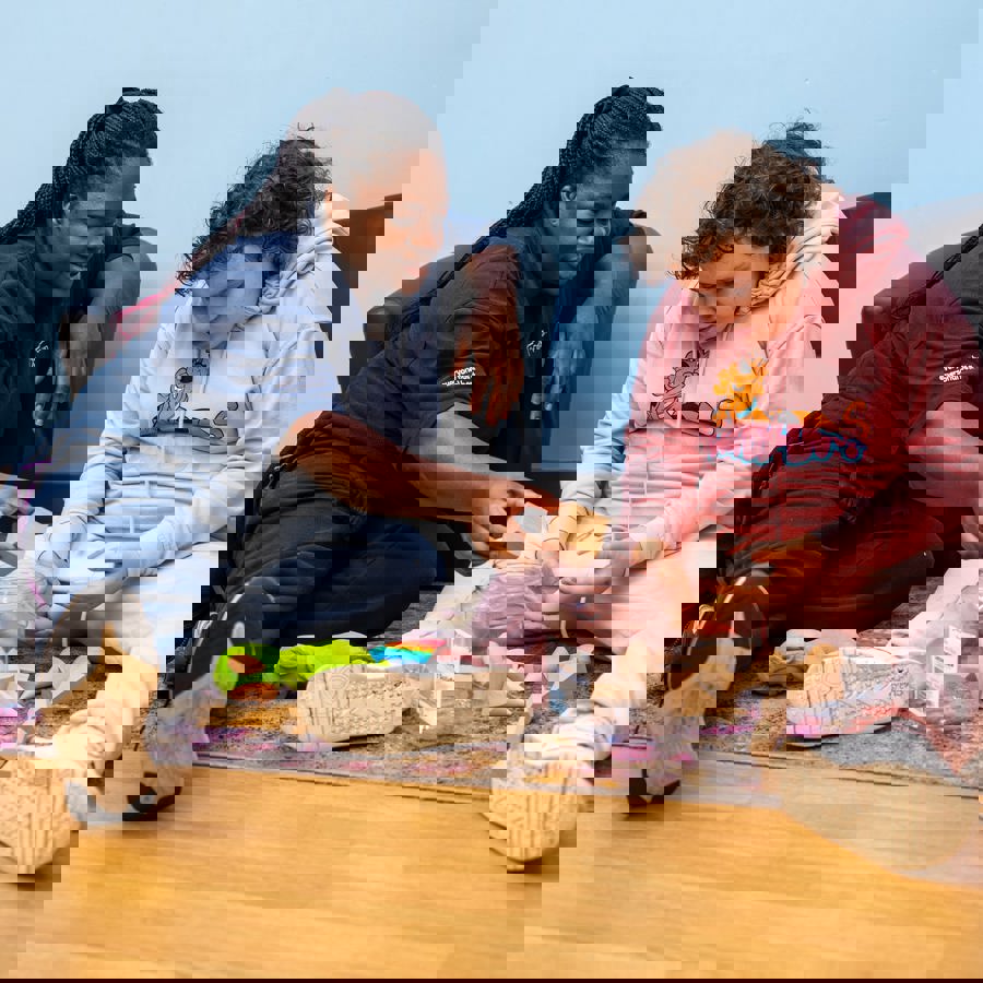 Carer sitting on the floor supporting a child as they explore and stack wooden blocks together.