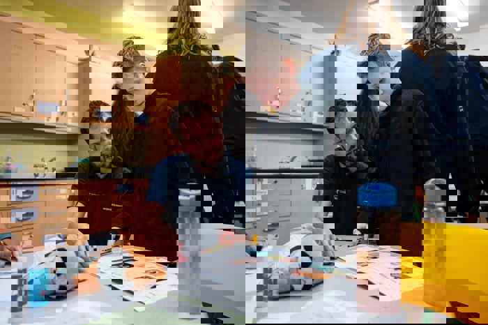 Adult supporting a child at a classroom table, pointing to a worksheet while the child follows along with a pencil.